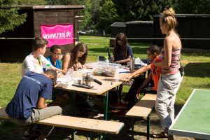 Junge Menschen sitzen vor einigen Holzhütten auf einer Wiese an einem Holztisch und schreiben etwas auf Zettel