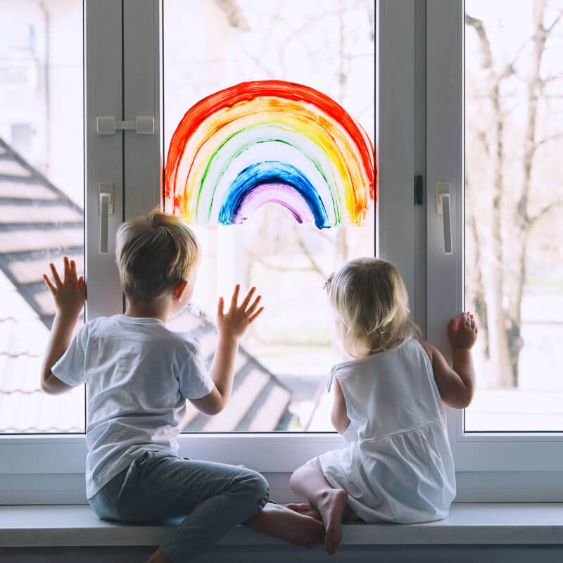 Zwei Kinder sitzen auf einer Fensterbank und schauen durch das Fenster. Auf diesem ist ein Regenbogen gemalt