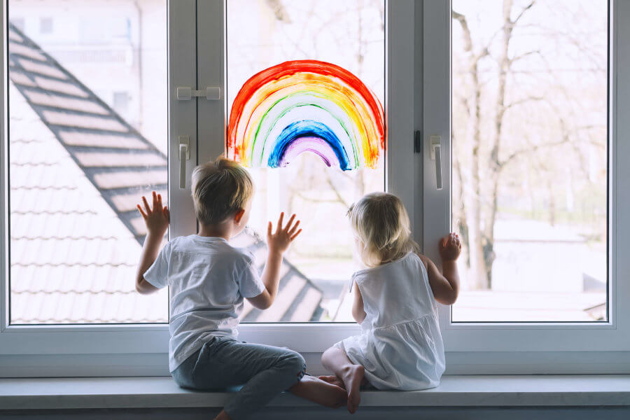 Zwei Kinder sitzen auf einer Fensterbank und schauen durch das Fenster. Auf diesem ist ein Regenbogen gemalt