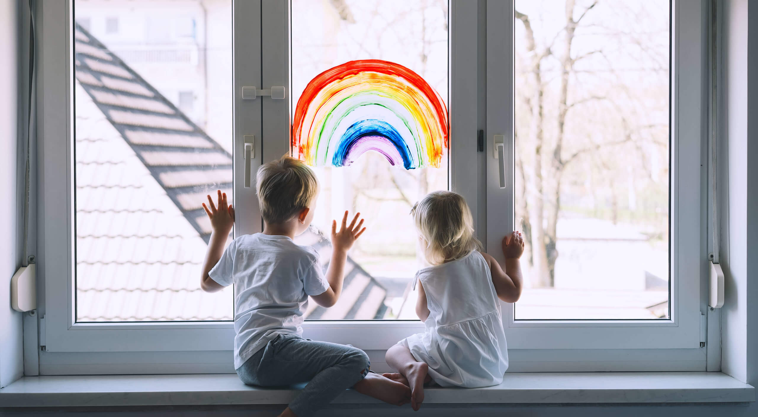 Zwei Kinder sitzen auf einer Fensterbank und schauen durch das Fenster. Auf diesem ist ein Regenbogen gemalt
