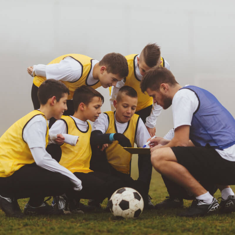 Ein Trainer zeigt 5 Jungen beim Fußballtrainig Dinge auf einem Klemmbrett