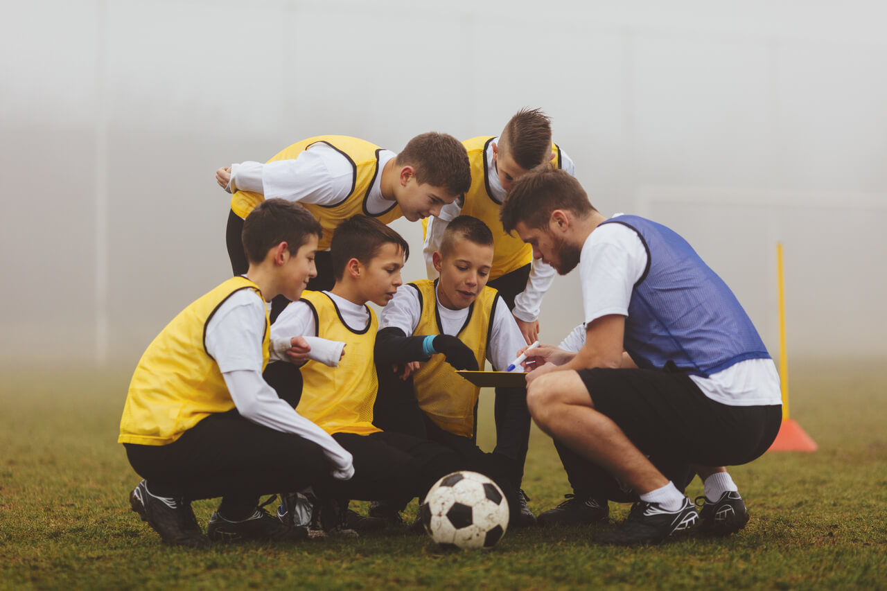 Ein Trainer zeigt 5 Jungen beim Fußballtrainig Dinge auf einem Klemmbrett