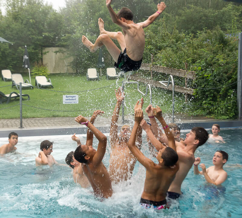 Gruppe von Jungs in einem Schwimmbecken. Einer wird in die Luft geworfen