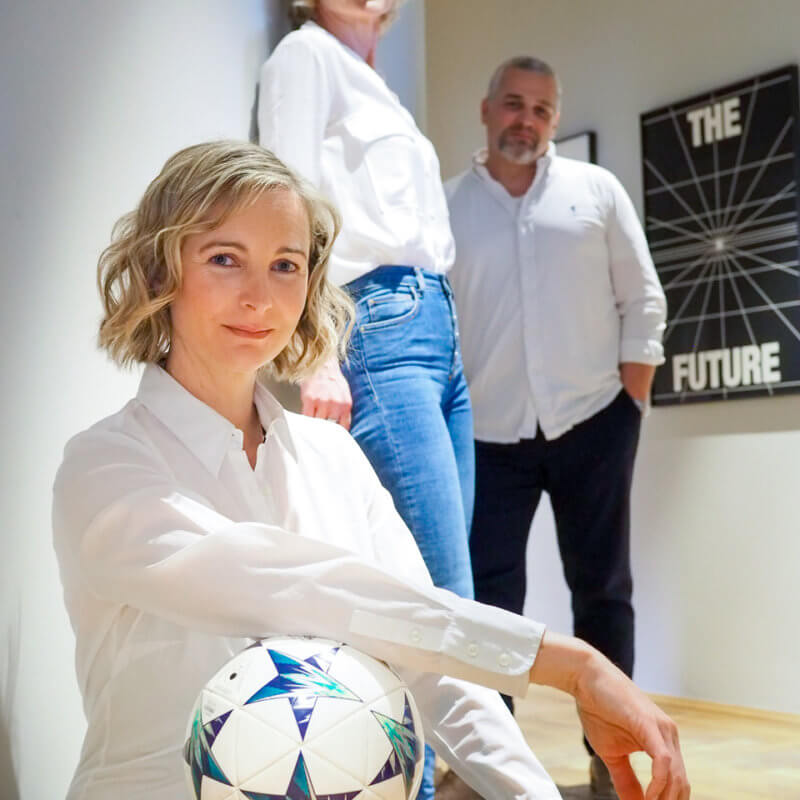 Drei Personen in einem Büro. Eine hält einen Fußball unter dem Arm