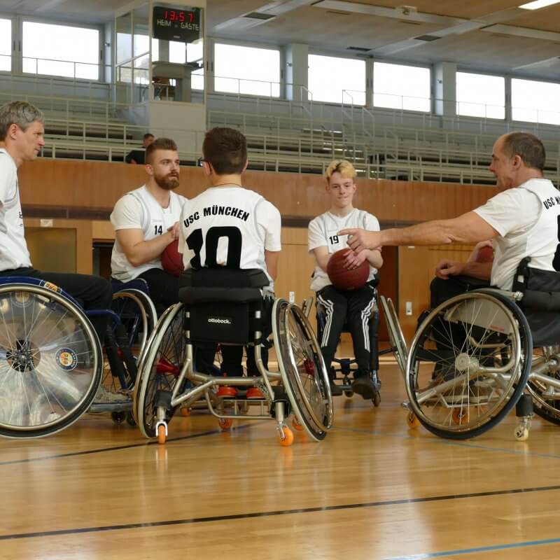 Sechs Rollstuhlfahrer in einer Turnhalle. Sie tragen ein Trikot und haben einen Basketball auf dem Schoß