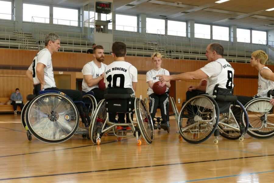 Sechs Rollstuhlfahrer in einer Turnhalle. Sie tragen ein Trikot und haben einen Basketball auf dem Schoß