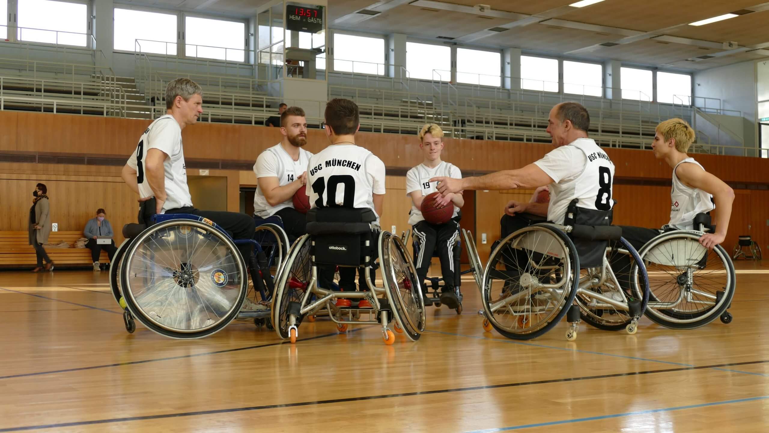 Sechs Rollstuhlfahrer in einer Turnhalle. Sie tragen ein Trikot und haben einen Basketball auf dem Schoß