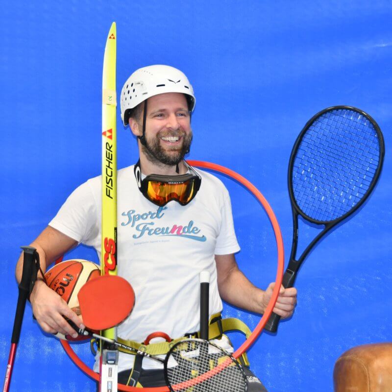 Mann mittleren Alters trägt ganz viele Sportgeräte wie Tennisschläger, Tischtennisschläger, einen Reifen um den Hals, Basketball, Badmintonschläger