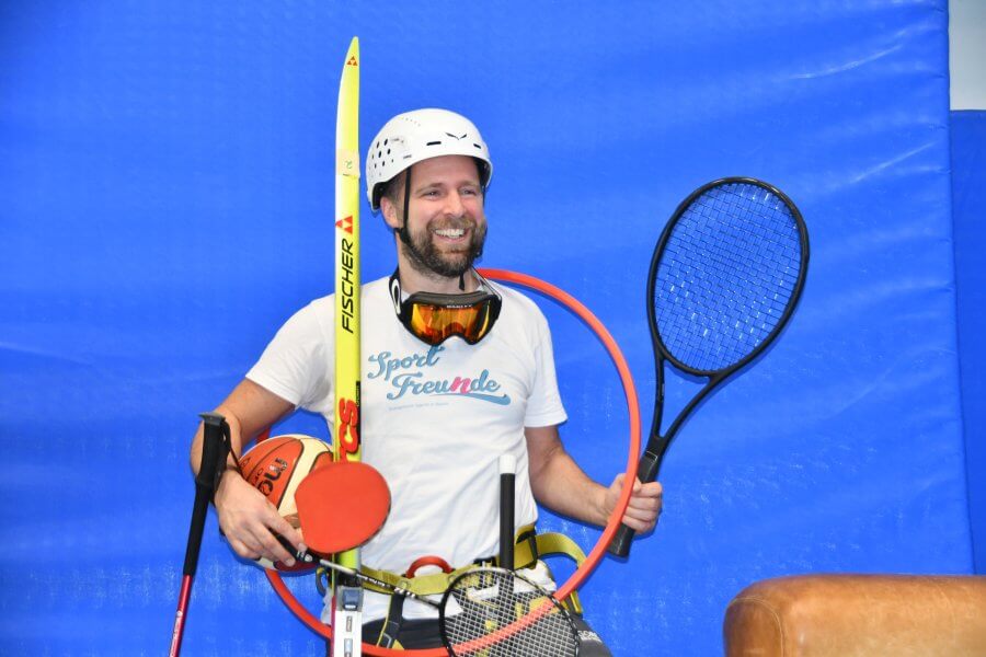 Mann mittleren Alters trägt ganz viele Sportgeräte wie Tennisschläger, Tischtennisschläger, einen Reifen um den Hals, Basketball, Badmintonschläger