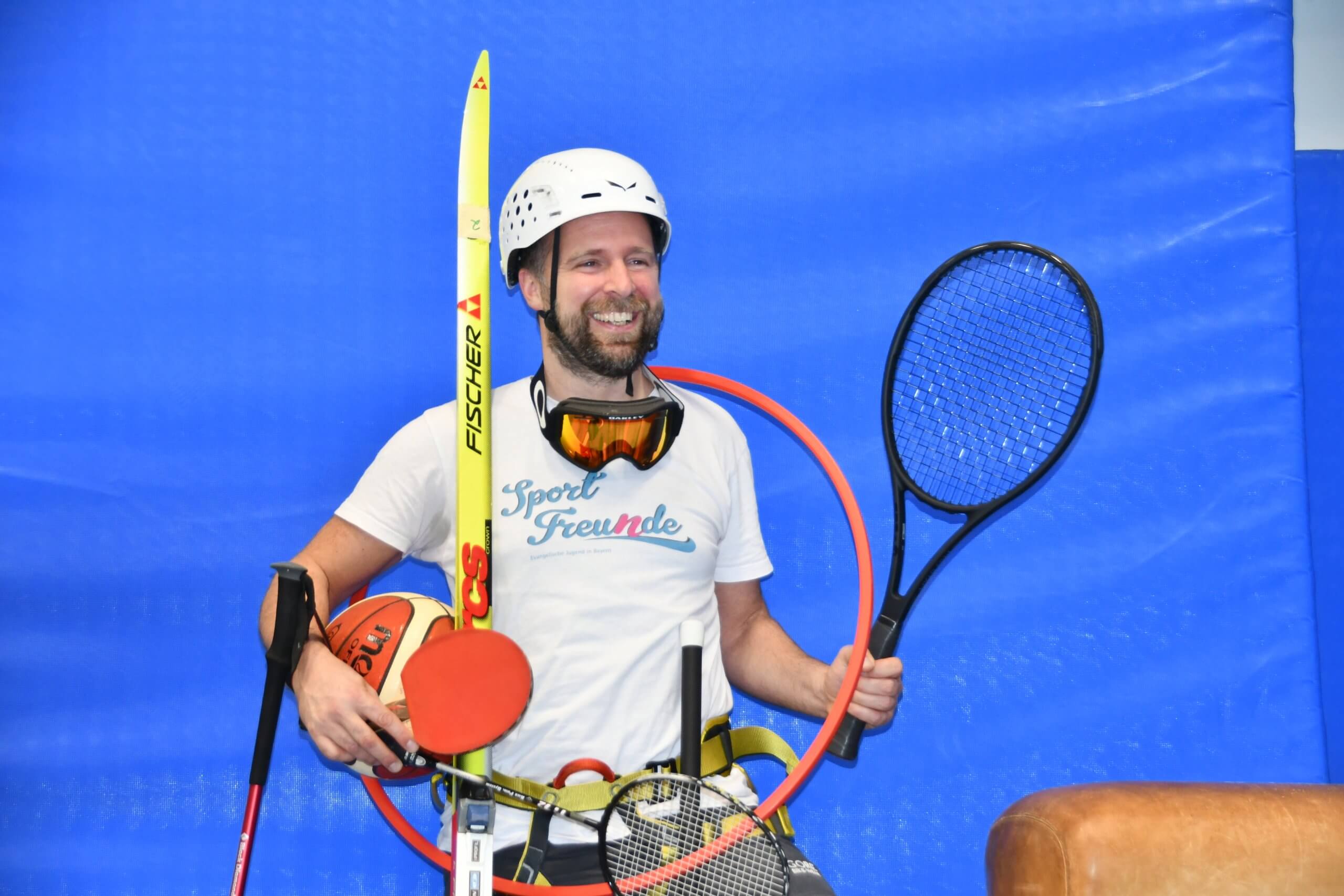Mann mittleren Alters trägt ganz viele Sportgeräte wie Tennisschläger, Tischtennisschläger, einen Reifen um den Hals, Basketball, Badmintonschläger