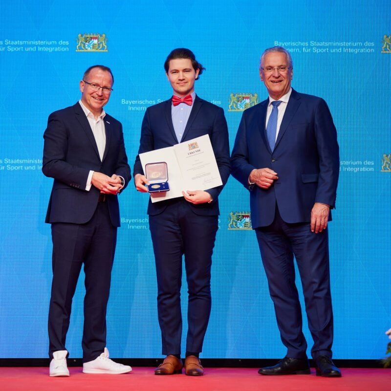 Drei Männer im Anzug. Einer hält eine Ehrenmedallie und Urkunde