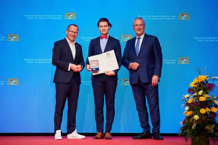 Drei Männer im Anzug. Einer hält eine Ehrenmedallie und Urkunde