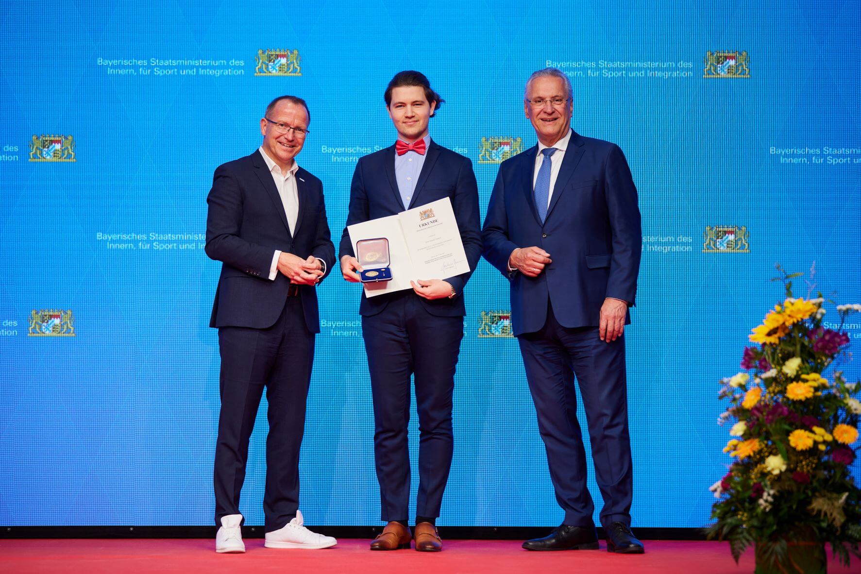 Drei Männer im Anzug. Einer hält eine Ehrenmedallie und Urkunde