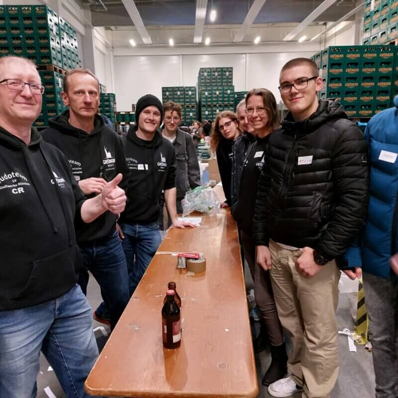 Gruppe von Erwachsenen stehen an einem Tisch. Im Hintergrund viele Paletten mit Getränkekisten