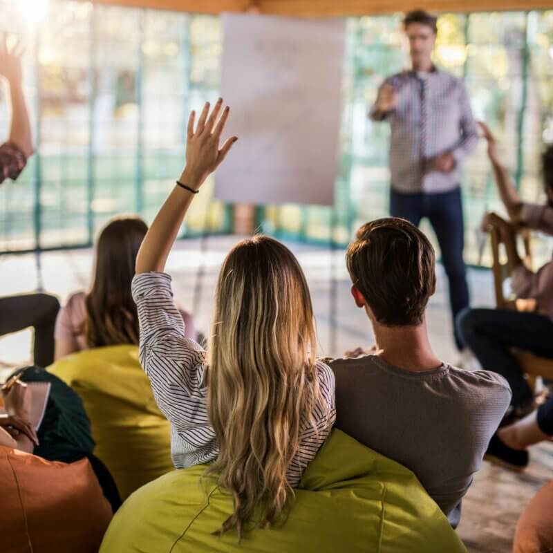 Eine Person steht an einem Flipchart. Andere Personen sitzen davor und melden sich