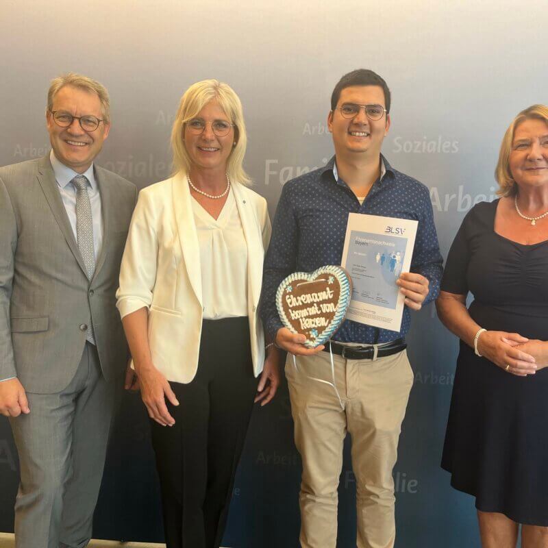 Zwei Männer und zwei Frauen vor einer Werbewand. Ein Mann hält ein Lebkuchenherz und eine Urkunde in der Hand