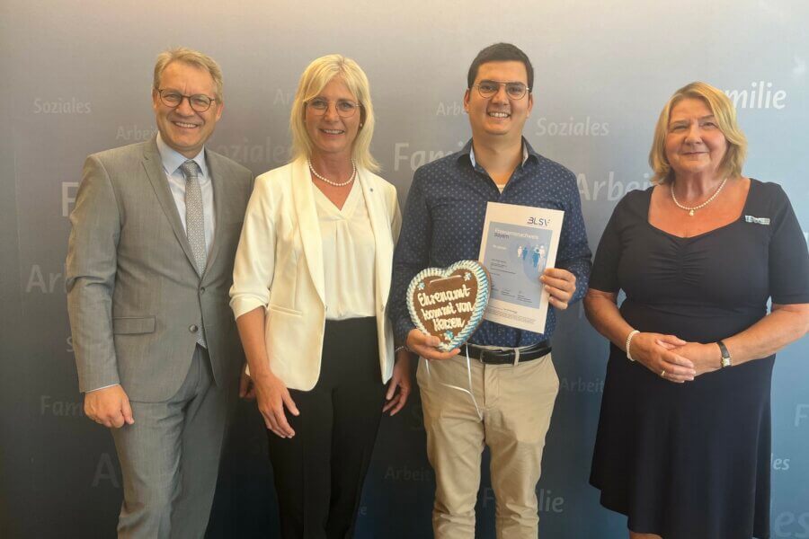 Zwei Männer und zwei Frauen vor einer Werbewand. Ein Mann hält ein Lebkuchenherz und eine Urkunde in der Hand