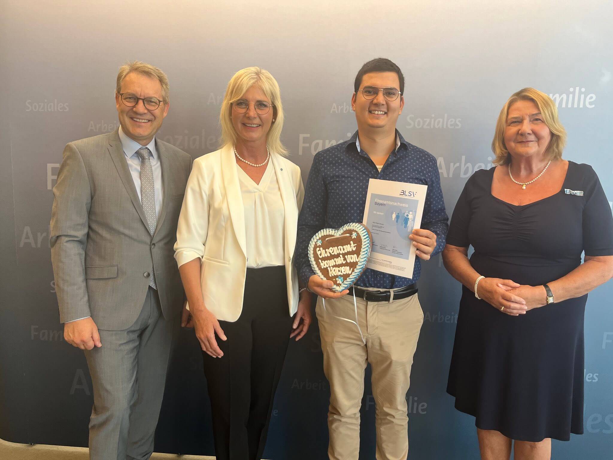 Zwei Männer und zwei Frauen vor einer Werbewand. Ein Mann hält ein Lebkuchenherz und eine Urkunde in der Hand