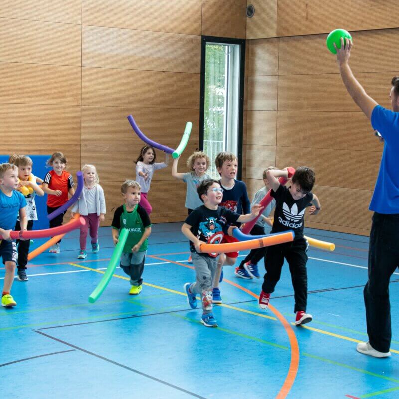 Kinder in Turnhalle halten jeweils eine Poolnoodle. Ein Mann hält einen Ball in die Luft