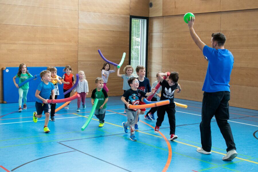 Kinder in Turnhalle halten jeweils eine Poolnoodle. Ein Mann hält einen Ball in die Luft