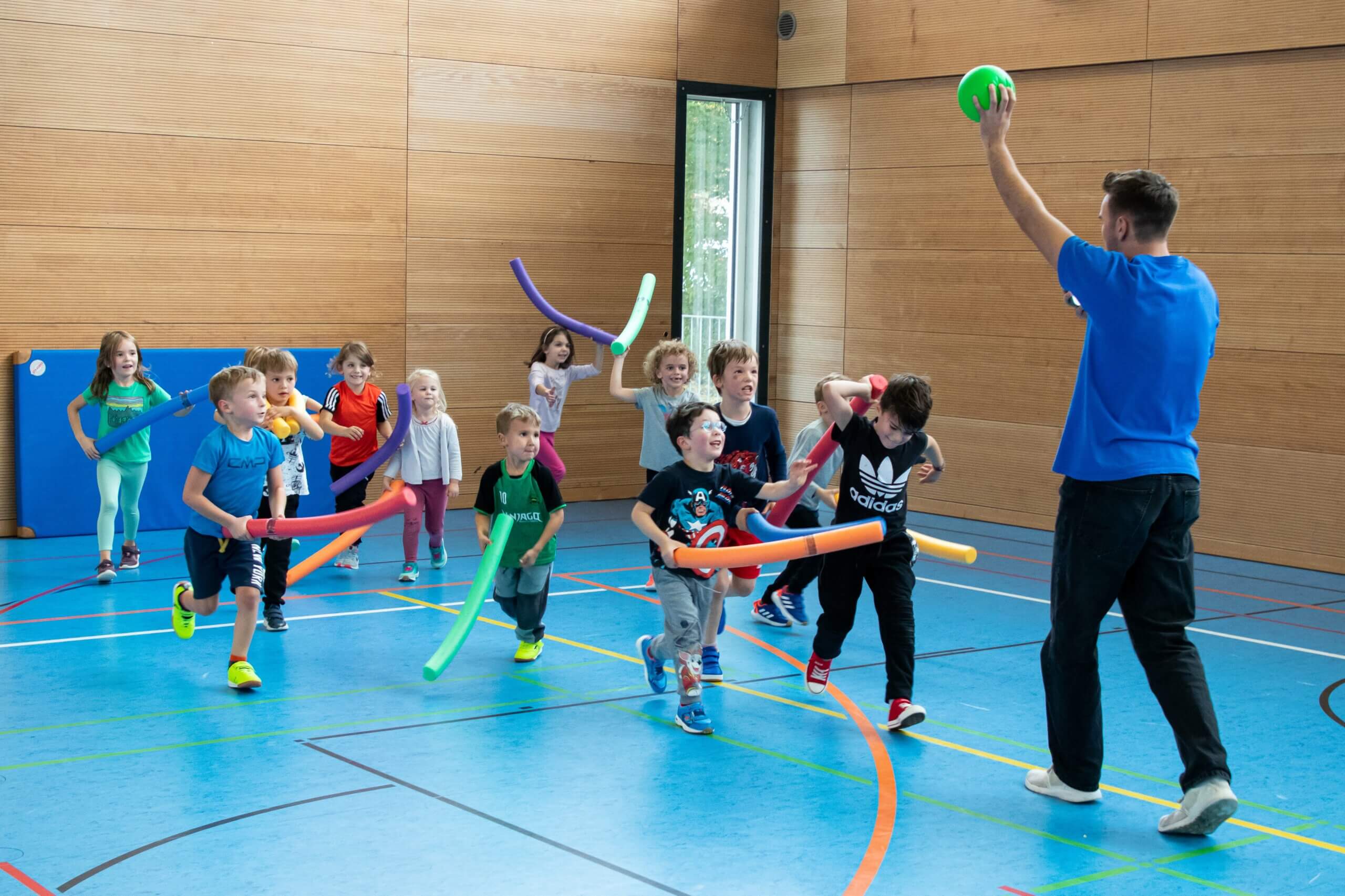 Kinder in Turnhalle halten jeweils eine Poolnoodle. Ein Mann hält einen Ball in die Luft