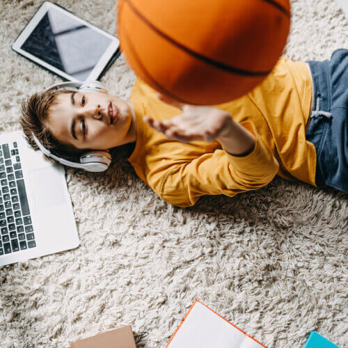 Ein Junge liegt auf dem Rücken und wirft einen Basketball in die Luft. Er trägt Kopfhörer. Neben ihm liegen ein Laptop und ein Tablet