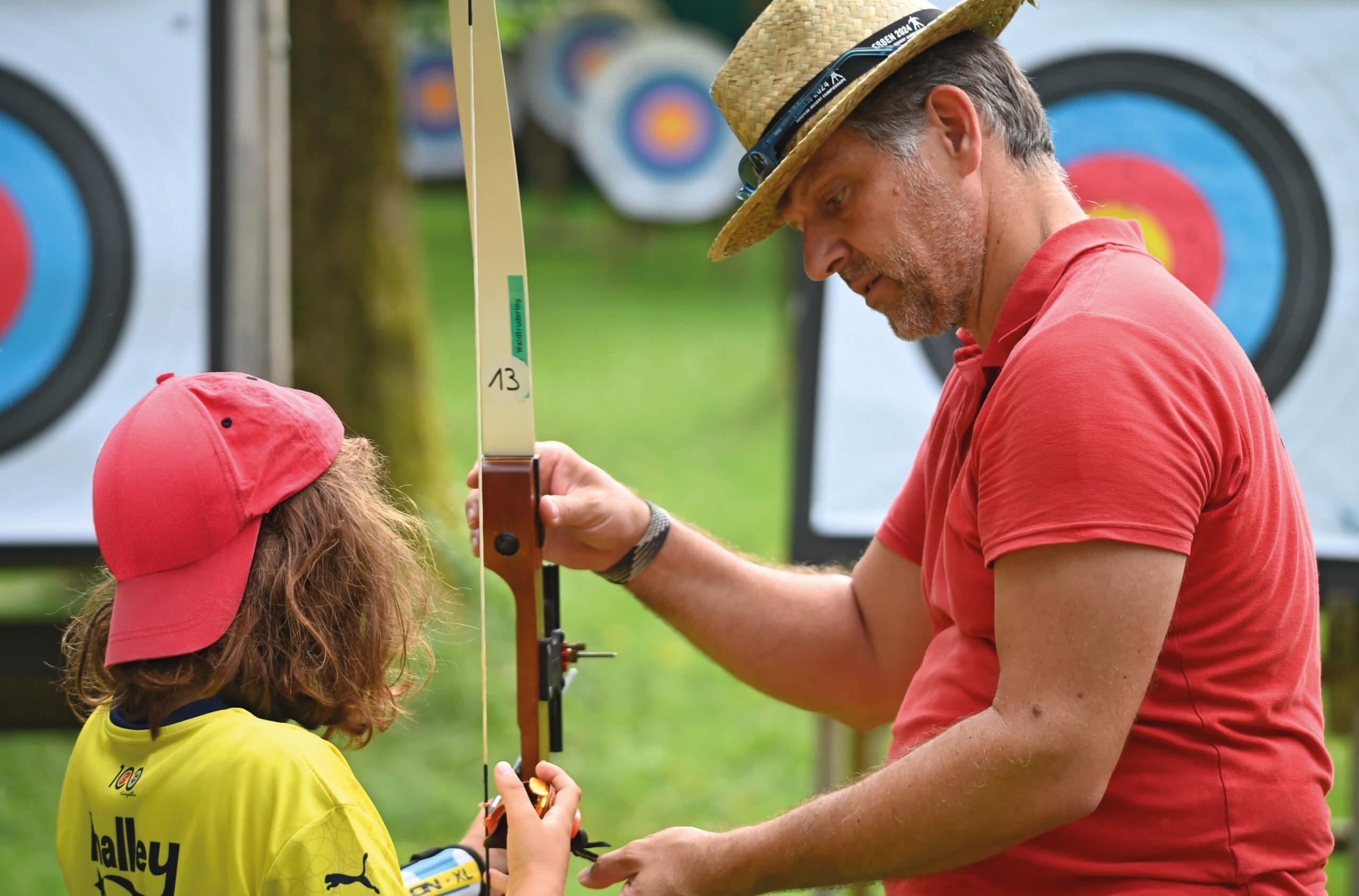 Ein Mann mit Strohhut hilft einem Kind beim Bogenschießen