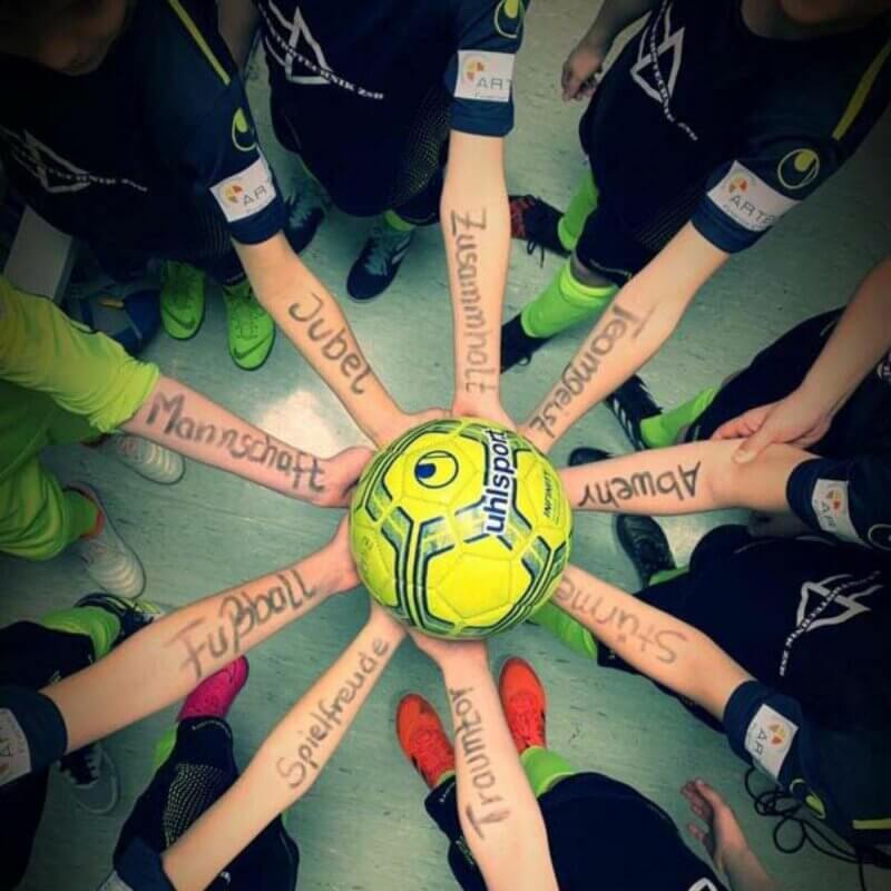 Acht Kinder halten einen einzigen Ball Hoch. Auf ihren Unterarmen sind Worte wie Jubel, Mannschaft oder Spielfreude geschrieben