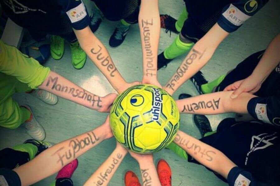 Acht Kinder halten einen einzigen Ball Hoch. Auf ihren Unterarmen sind Worte wie Jubel, Mannschaft oder Spielfreude geschrieben
