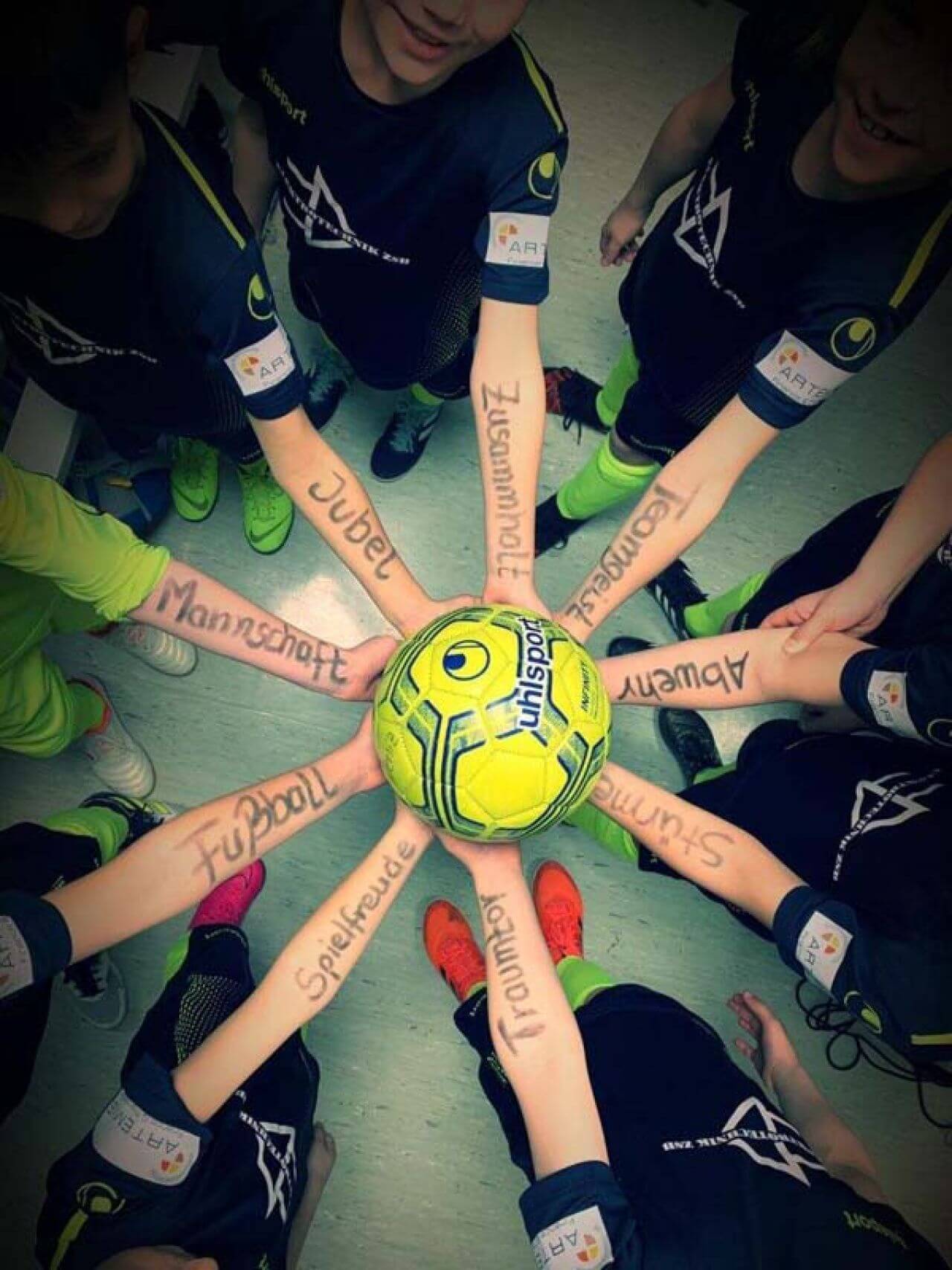 Acht Kinder halten einen einzigen Ball Hoch. Auf ihren Unterarmen sind Worte wie Jubel, Mannschaft oder Spielfreude geschrieben