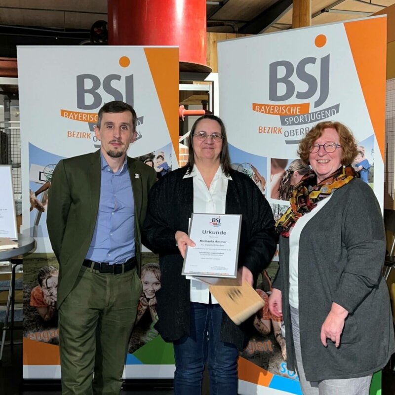 Ein Mann und zwei Frauen stehen vor einer Werbefläche der BSJ. Die Frau in der Mitte hält eine Urkunde vor der Brust