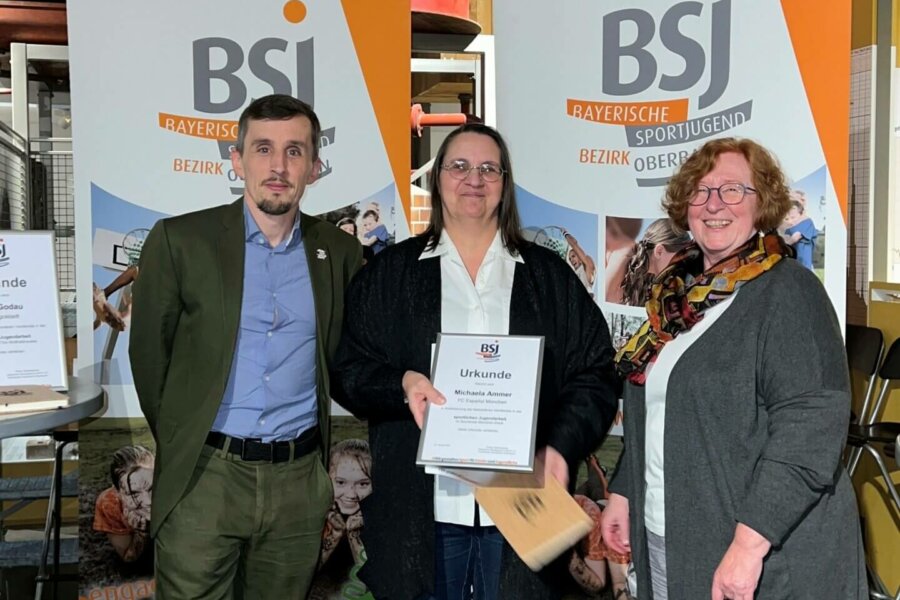 Ein Mann und zwei Frauen stehen vor einer Werbefläche der BSJ. Die Frau in der Mitte hält eine Urkunde vor der Brust