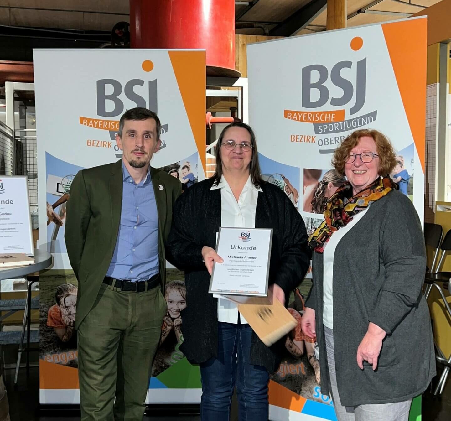 Ein Mann und zwei Frauen stehen vor einer Werbefläche der BSJ. Die Frau in der Mitte hält eine Urkunde vor der Brust