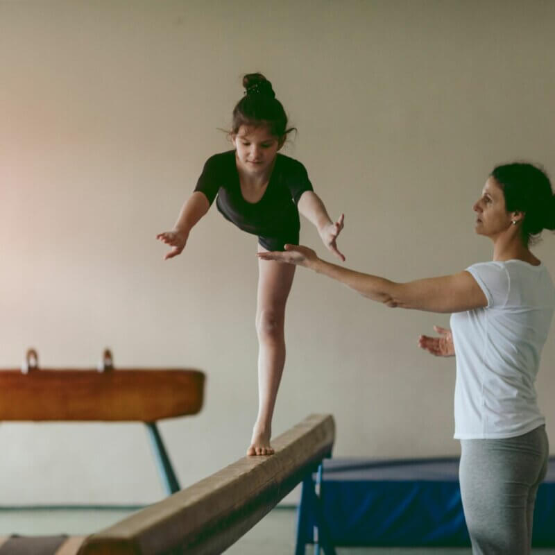 Ein Mädchen turnt auf einem Schwebebalken. Eine Frau passt auf, dass sie nicht fällt