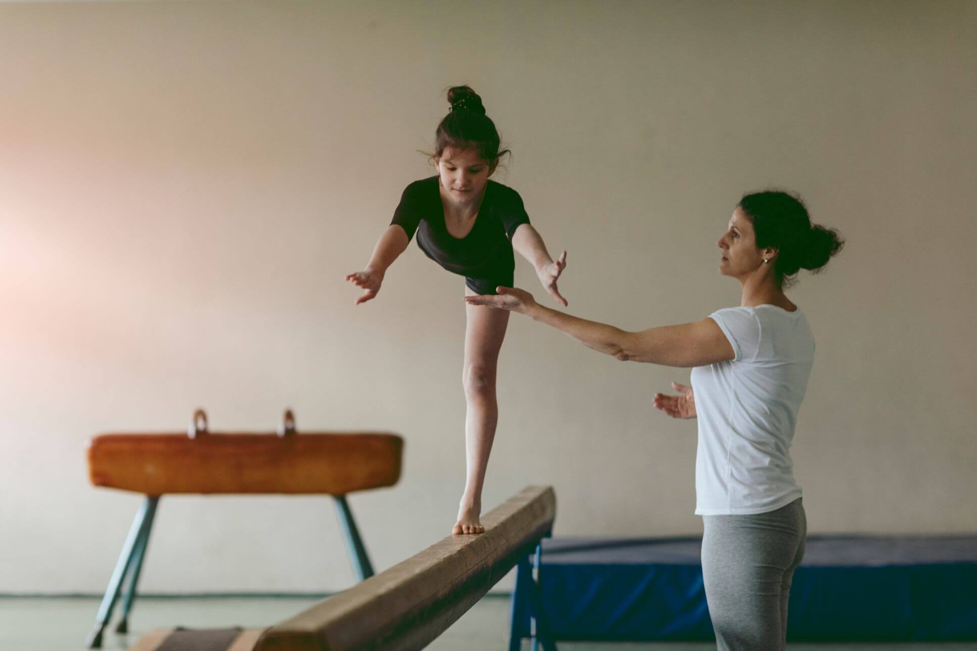 Ein Mädchen turnt auf einem Schwebebalken. Eine Frau passt auf, dass sie nicht fällt